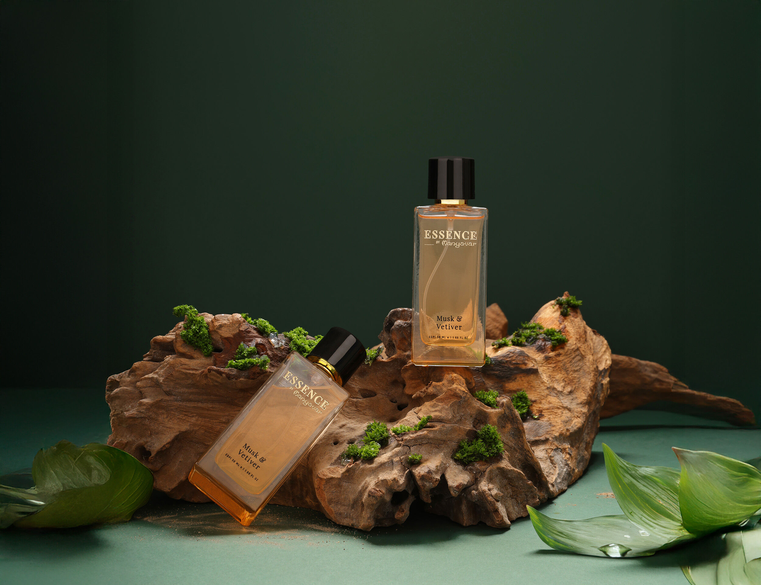Essence by Manyavar Musk & Vetiver perfumes displayed on natural driftwood with green moss and tropical leaves against a dark green background